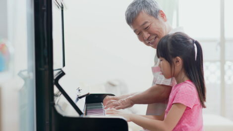 Elderly man smiles while playing piano with young girl.