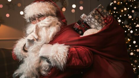 Smiling Santa with index finger to his lips, making a shushing gesture.