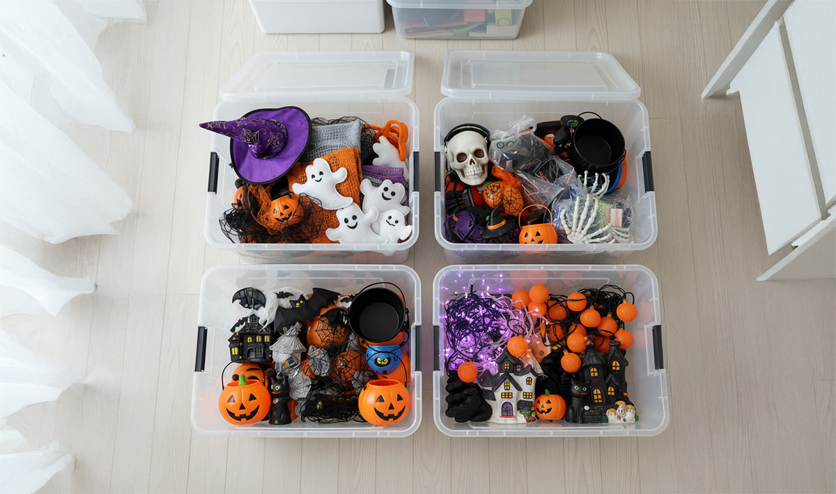 Four clear plastic storage totes filled with various Halloween decorations, including witch hats, ghost figures, pumpkin buckets, skulls, spiderwebs, and string lights, organized on a light-colored floor. Ideal for a Halloween decoration storage guide by MyPlace Self Storage.